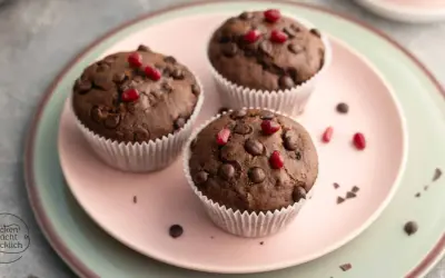 Drei saftige, vegane Muffins mit Schokostückchen und Granatapfelkernen auf einem rosa Teller.