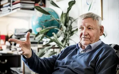 Äldre man med grått hår, liknar Ragnar Skanåker, gestikulerar med handen. Bakom honom syns en jordglob och växter.