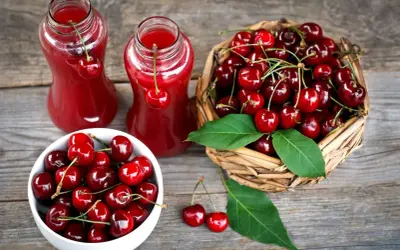 Dwa szklane dzbanki z rubinowym sokiem z czereśni, obok koszyk pełen świeżych owoców i miseczka z czereśniami.