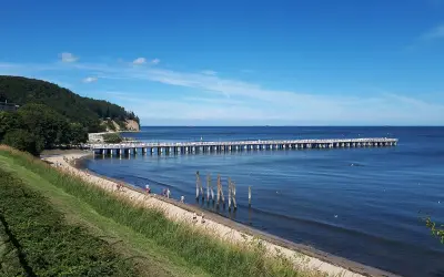 En lång pir sträcker sig ut i det blå havet vid Gdynia, Polen. En grön kulle med träd reser sig vid sidan av stranden där några människor promenerar.