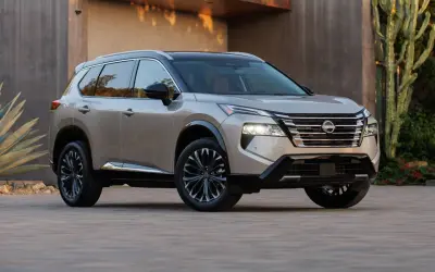 Elegancki, beżowy Nissan Rogue z czarnym dachem i przyciemnianymi felgami stoi przed nowoczesnym budynkiem, otoczony egzotyczną roślinnością.