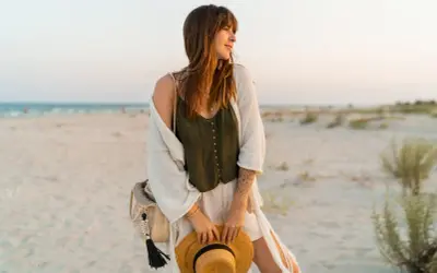 Kobieta w letnich stylizacjach boho jesień na plaży, z kapeluszem w dłoni i plecioną torbą.