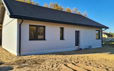Nowy dom z szarym dachem i białymi ścianami, otoczony piaskiem i jesiennymi drzewami. Ssz.