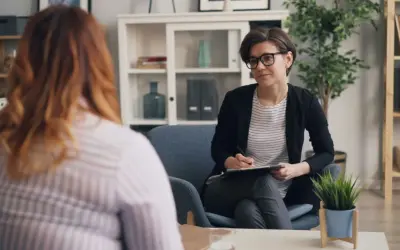Psychoterapeutka w okularach rozmawia z pacjentką, notując coś na kartce.