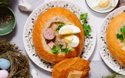 Pyszny żur w chlebie z kiełbasą i jajkiem, ozdobiony natką pietruszki. Na stole wielkanocne dekoracje: pisanki w gnieździe, zajączek.