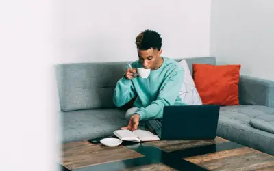 Ung man dricker koffeinhaltigt kaffe medan han läser och arbetar vid sin laptop i soffan.