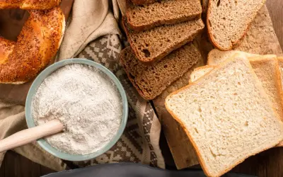 Mąka w misce, kromki chleba i obwarzanek. W tych produktach jest gluten, który jest podstawą wypieków.
