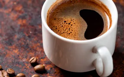 En vit kopp med mörkt, skummande kaffe. Kaffe koffein per 100 ml kan variera, men denna kopp ser stark ut. Kaffebönor ligger utspridda.