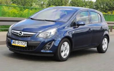 Granatowy Opel Corsa D jedzie po drodze, odbijając niebo i chmury.