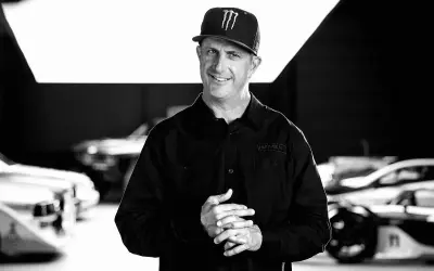 Ken Block, sonriente y con gorra, posa frente a coches de rally.