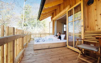 Frau liest im Bett in einer Holzhütte mit offener Tür und Blick auf Bäume.