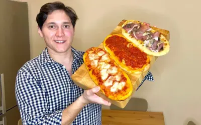 Uomo sorridente mostra tre lingue di pizza senza lievito: una margherita, una al pomodoro e una con prosciutto e funghi.