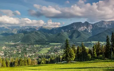 Zielona polana z widokiem na malownicze lasy w Tatrach, miasteczko i skaliste szczyty w tle.