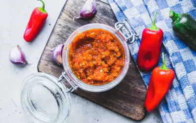 Domowy sos sriracha z papryczek chilli i czosnku w słoiku, otoczony świeżymi papryczkami i czosnkiem na desce.