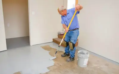Un homme en bleu peint le sol en béton avec un rouleau. Il porte des genouillères pour peindre le béton confortablement.