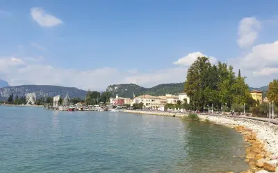 Il lungolago di Bardolino, con il lago azzurro, le barche, le case colorate e le montagne sullo sfondo. Una ruota panoramica si staglia all'orizzonte.