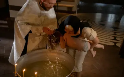 Kapłan w białej szacie błogosławi wodę, polewając nią głowę dziecka podczas chrztu.