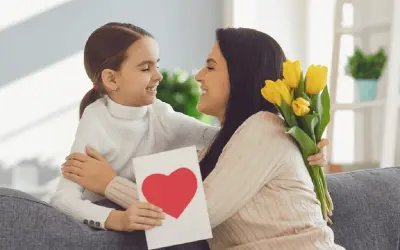 Uśmiechnięta dziewczynka wręcza mamie kartkę z sercem i bukiet żółtych tulipanów. Radosny Dzień Matki UK!