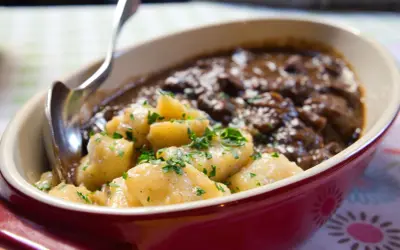 Gulasz z dzika z kluskami ziemniaczanymi, posypany natką pietruszki. Aromatyczne danie w bordowej misie, gotowe do podania.
