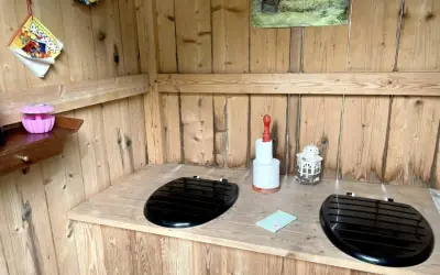 Två svarta sitsar på en träbänk i ett utedass. En rulle toalettpapper och en lykta står på bänken. En bild med hästar hänger på väggen. Här finns utedass regler.