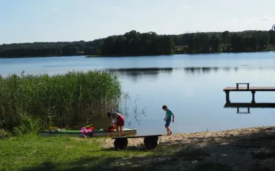 Dzieci szykują kajak nad spokojnym jeziorem w gminie Morąg. W tle las i drewniane molo.