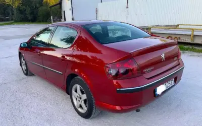Błyszczący czerwony Peugeot 407 2.0 HDI z przyciemnianymi szybami stoi na żwirowym podjeździe.