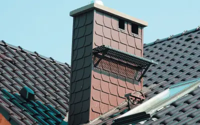 Czapka na komin dymowy chroni przed deszczem i ptakami. Widać ją na dachu z brązowej dachówki.