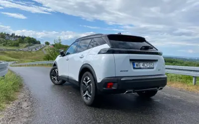 Biały Peugeot 2008 GT na mokrej drodze, z widokiem na krętą trasę i zielone wzgórza.