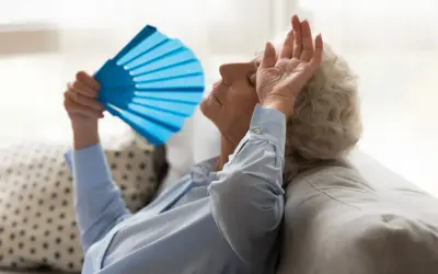 Starsza kobieta zmagająca się z uderzeniami gorąca, wachlująca się niebieskim wachlarzem. Może to być związane z menopauzą, którą leczy fezolinetant.