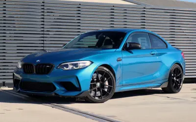 Błękitne BMW M2 Competition z czarnymi felgami i detalami, zaparkowane przed nowoczesną, szarą ścianą.