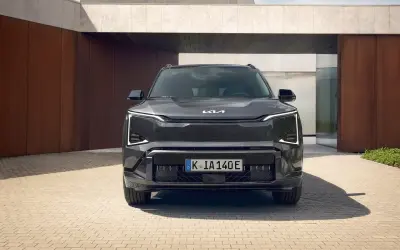Nowoczesny, ciemnoszary Kia EV5 z charakterystycznymi światłami LED stoi przed minimalistycznym budynkiem z elementami rdzy i betonu.