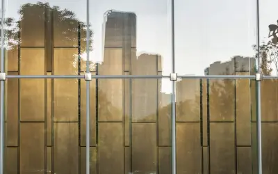 Reflejos dorados de árboles y edificios en un **cristal blindado** moderno, con estructura metálica visible.