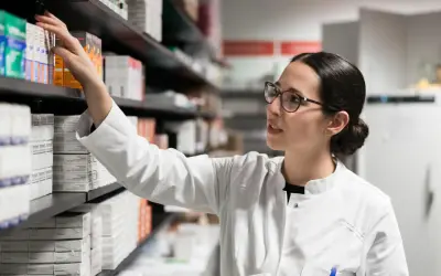 Farmaceutka w białym kitlu sięga po lek, być może pentoksyfilinę, z aptecznej półki pełnej opakowań.