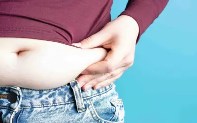 Mano che stringe la pancia, evidenziando gonfiore e possibile ritenzione idrica pancia. Jeans a vita bassa e maglia bordeaux.