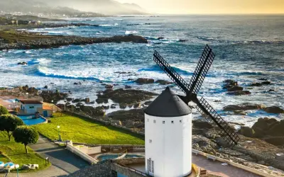Un muiño do vento blanco con aspas negras se alza junto a un mar azul y rocoso. El paisaje costero es agreste y hermoso.