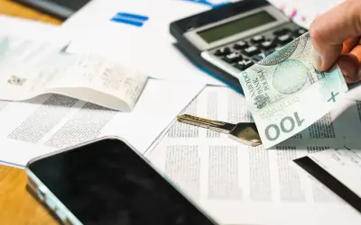 Dłoń trzyma banknot 100 zł nad dokumentami, kluczem i telefonem. To potwierdzenie odbioru gotówki.