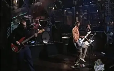 Zespół gra na scenie, muzycy chcą nauczyć się latać, a basista w czerni i gitarzyści w kolorowych koszulach tworzą energię.