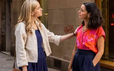 Zwei Frauen unterhalten sich auf einer Straße. Die eine trägt ein blaues Kleid und eine weiße Jacke, die andere ein buntes Hemd. Szene aus "The Diplomat" Serie ZDF.