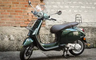 Elegancki, ciemnozielony skuter Piaggio z brązowym siedzeniem i chromowanymi dodatkami stoi na tle ceglanej ściany.