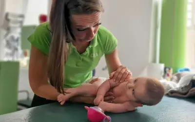 Fizjoterapeutka pracuje z niemowlęciem, pomagając mu w walce z **hipertonią**. Dziecko leży na zielonym stole, a kobieta delikatnie dotyka jego rączek.