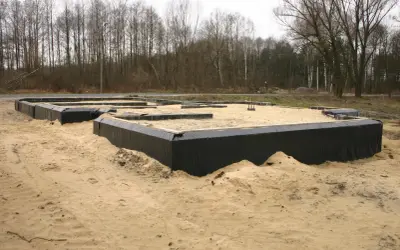 Fundamenty domu w piasku, otoczone czarną folią. W tle las. Wkrótce powstanie tu chudy beton.