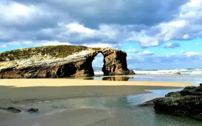 Arco natural en las playas de Galicia, con arena dorada, mar azul y cielo nublado.