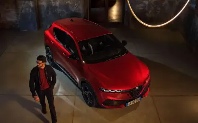 Elegancki mężczyzna w okularach obok lśniącego, czerwonego Alfa Romeo Tonale. Stylowy duet w industrialnym wnętrzu.