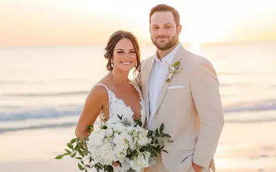 Groom in a tan suit, perfect for what to wear to beach wedding male, smiles with bride holding bouquet at sunset.