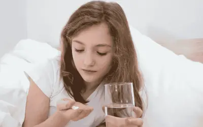 Dziewczynka w łóżku trzyma w dłoni tabletki i szklankę wody. Uważnie patrzy na białe tabletki, które mogą być przeterminowany paracetamol dla dzieci.