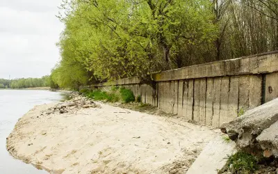 Betonowa opaska na rzece chroni brzeg przed erozją. Wiosenne drzewa tworzą zieloną ścianę nad wodą.