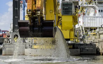 Gelbe Baggerschiff-Arme entleeren Wasser und Sedimente ins Meer. Ein riesiger Eimer schaufelt Material aus dem Wasser.