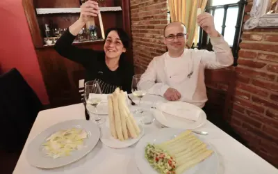 Pareja sonríe mostrando espárragos blancos, uno de ellos en un plato con ensalada y el otro en un manojo atado.