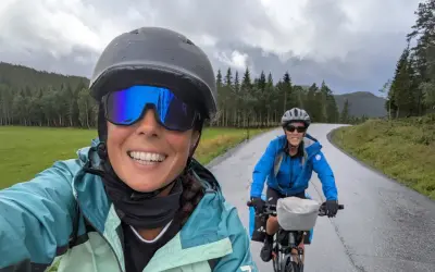 Dos ciclistas disfrutan de la ruta EuroVelo 6, con un paisaje verde y montañoso bajo un cielo nublado.