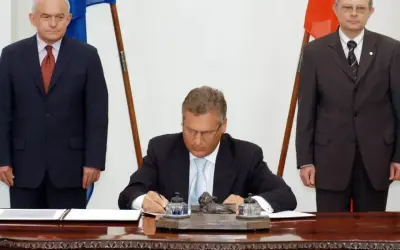 Uroczyste podpisanie dokumentów, symboliczny moment, kiedy Polska weszła do Unii. Trzech mężczyzn w garniturach, flagi Polski i UE.